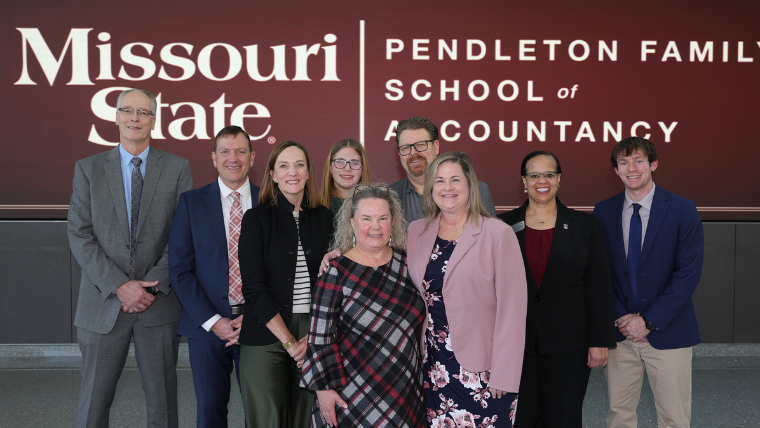 A diverse group of ten individuals, including women, men, and a young girl, smiles at the camera, standing in two rows. The background is a dark red wall with white text reading "Missouri State" on the left and "PENDLETON FAMILY SCHOOL of ACCOUNTANCY" on the right. The group wears business casual and professional attire, including suits, blazers, a plaid top, and a floral dress.