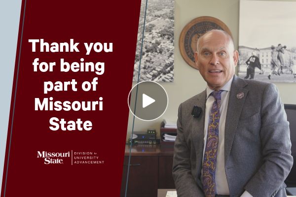 A video thumbnail split vertically: the left third has a maroon background with white text "Thank you for being part of Missouri State" and the Missouri State Advancement logo; the right two-thirds shows a smiling bald man in a grey suit and purple-gold paisley tie, wearing a lapel mic, looking directly at the camera from an office setting with black-and-white photos and a university seal behind him. A large white play button overlays the center.